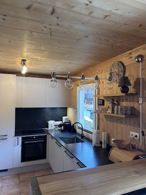 Une cuisine moderne avec des meubles en bois, un plan de travail noir et un four encastré. Des luminaires suspendus et une fenêtre laissent entrer la lumière naturelle.