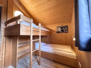 Une pièce de cabane en bois avec des lits superposés, une échelle, une petite fenêtre et un luminaire fixé au mur.