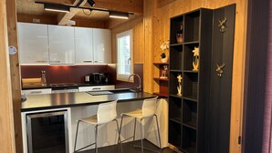 Cocina compacta con armarios blancos, encimera negra y horno empotrado. Hay dos taburetes blancos y un refrigerador pequeño.