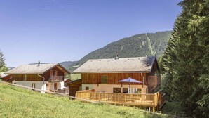 Una cabaña de madera con balcón y terraza, rodeada por un bosque y montañas.