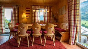 Une salle à manger en bois, avec une table et des chaises, des rideaux à carreaux et une vue sur l’extérieur.