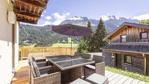 Eine Holzterrasse mit Korbmöbeln und einem Glastisch, mit Blick auf eine Berglandschaft.
