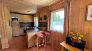 Interior d'una cabana de fusta amb cuina, tamborets d'abarco, un rellotge i una finestra amb cortines.
