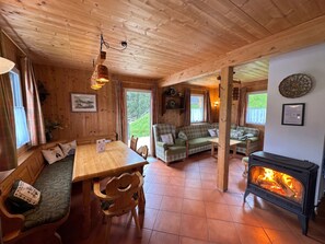 A wooden cabin interior with a dining table, benches, a sofa, a fireplace, and a view through a door.