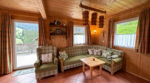 A cozy cabin interior with a green plaid sofa, two armchairs, a wooden table, and a balcony with a view of the forest.