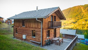 Eine Holzhütte mit einem Balkon, einer Terrasse mit Stühlen und Blick auf ein bewaldetes Tal.
