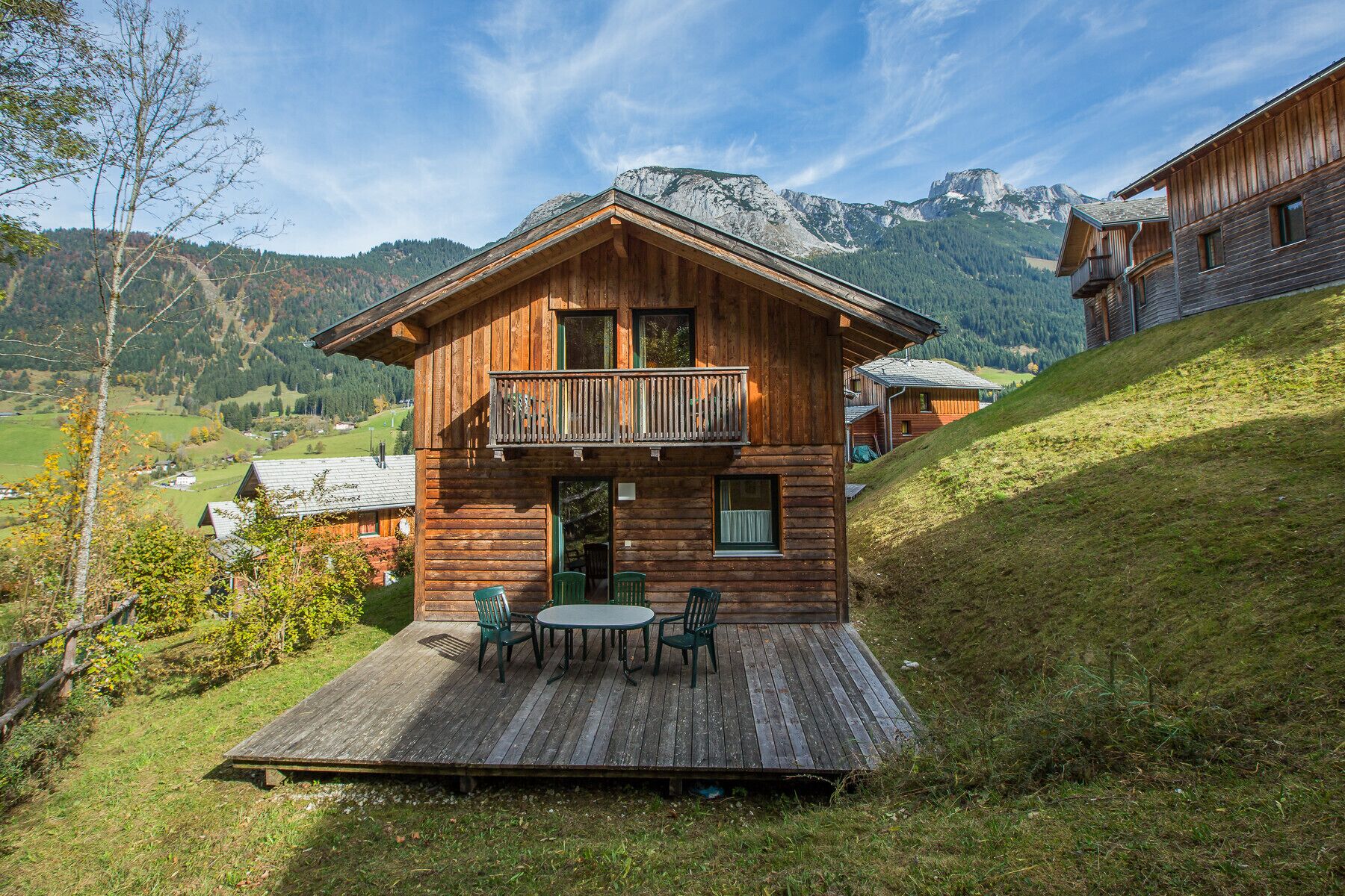 A wooden chalet with a balcony, a deck with outdoor furniture, and a mountainous backdrop.