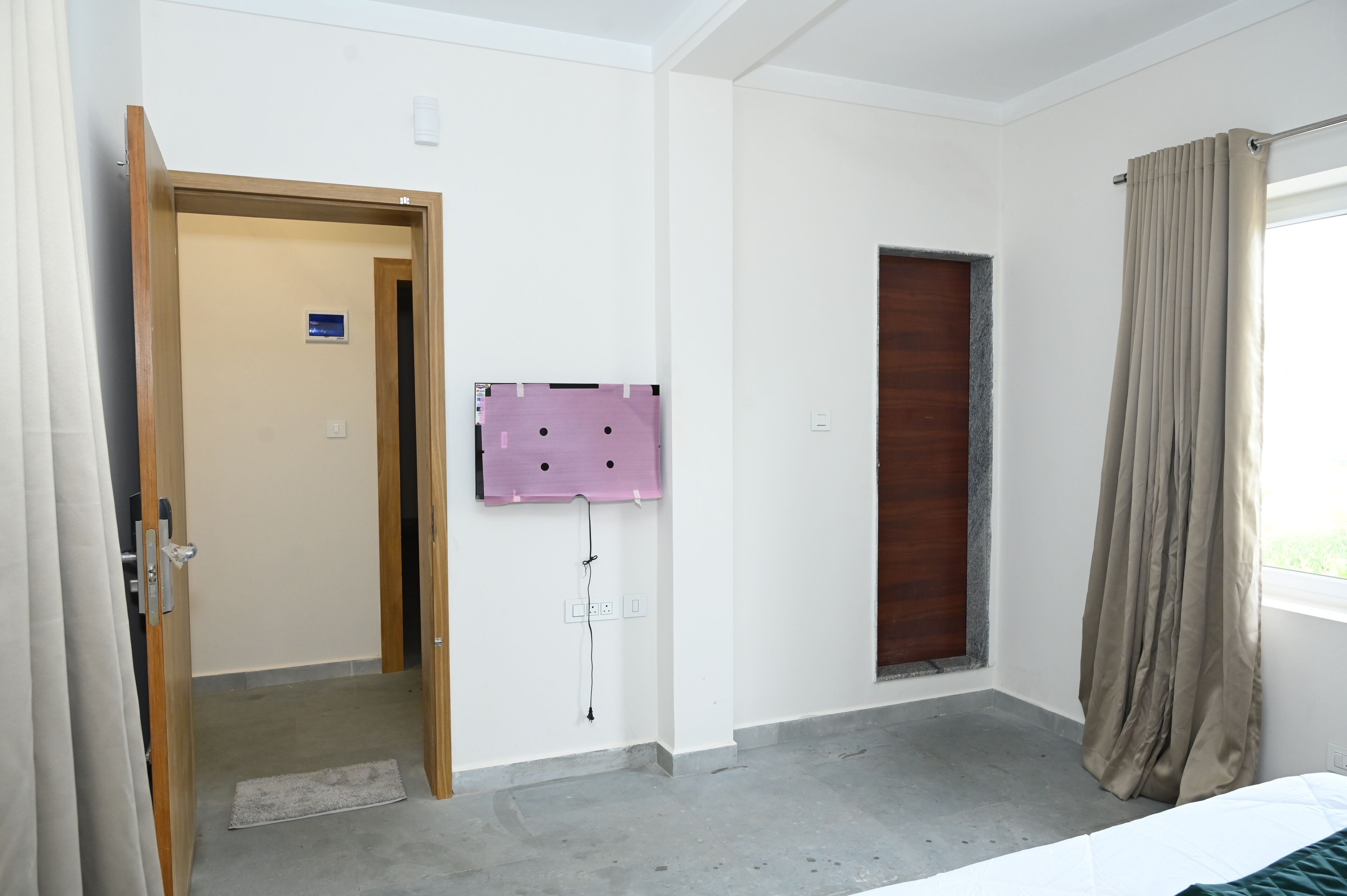 A hotel room with a wooden door, a small window with curtains, a wall-mounted air conditioning unit, and a visible door handle.