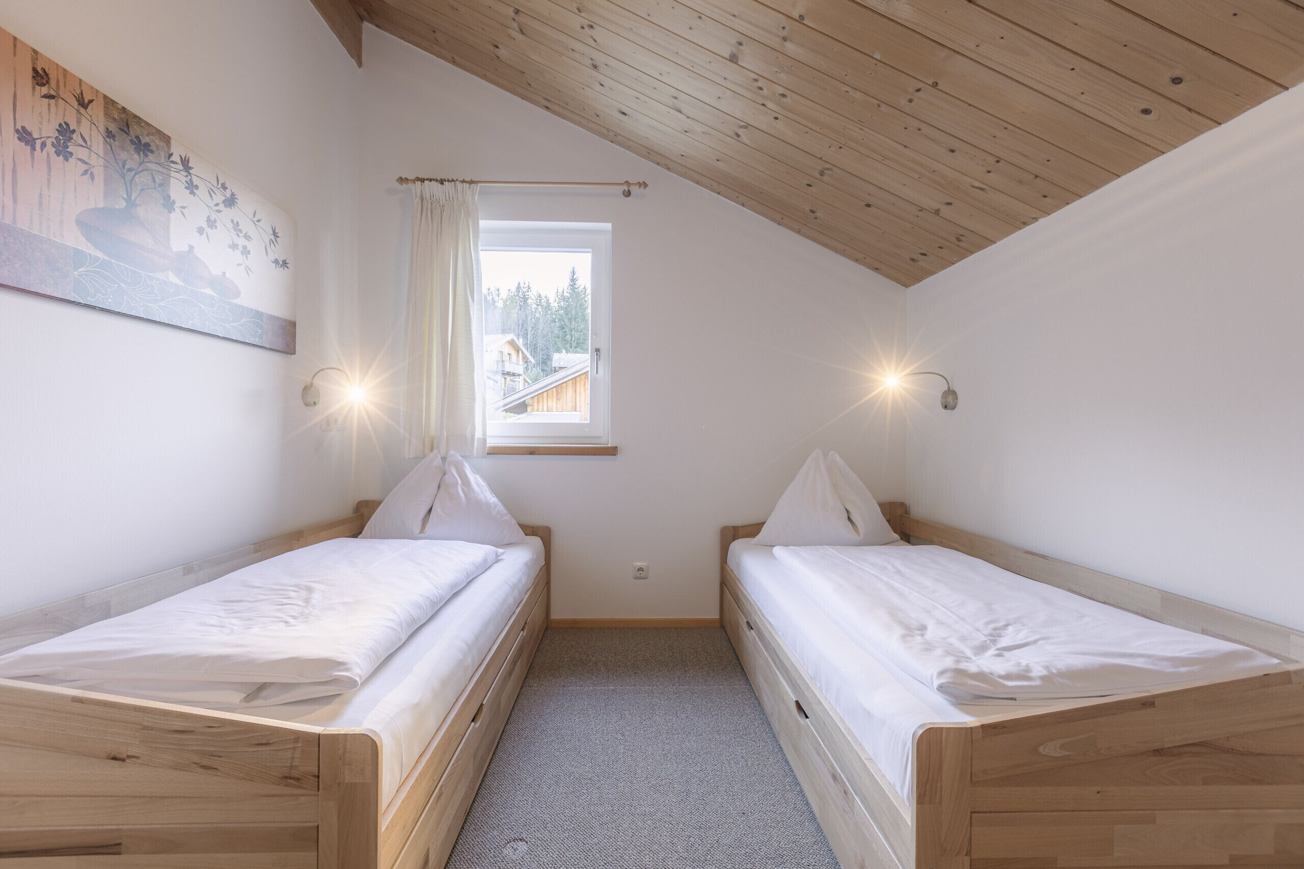 A small, well-lit room with two wooden beds, a window with a view of a house, and a painting on the wall.