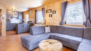 L’intérieur d’une maison en rondins moderne, avec un canapé d’angle gris, un plancher en bois, un coin cuisine et de grandes fenêtres dotées de rideaux.