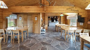 Interior de una cabaña de madera con suelo de piedra, mesas y sillas de madera, chimenea central y un letrero que indica un lugar de vacaciones en un pueblo.