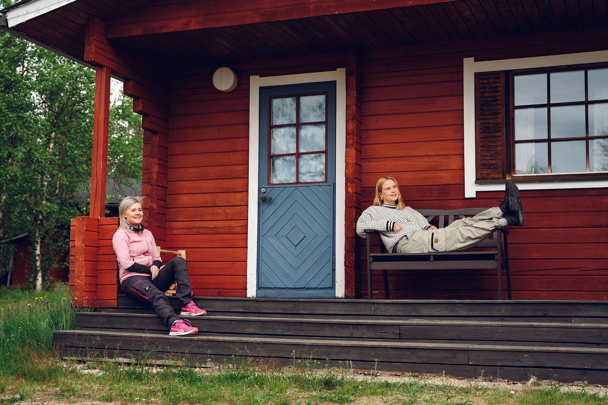 Deux personnes sont assises sur un banc devant une cabane en bois avec une porte et des fenêtres bleues.