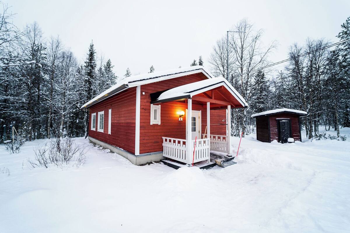 Una cabaña roja con un porche y un cobertizo pequeño adosado, rodeada de árboles cubiertos de nieve.