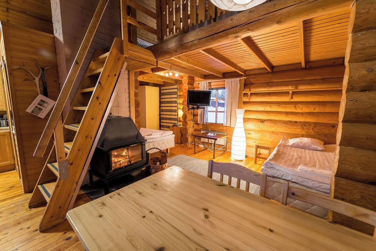 Interior acogedor de una cabaña de troncos con escalera de madera, chimenea, televisor y cama.