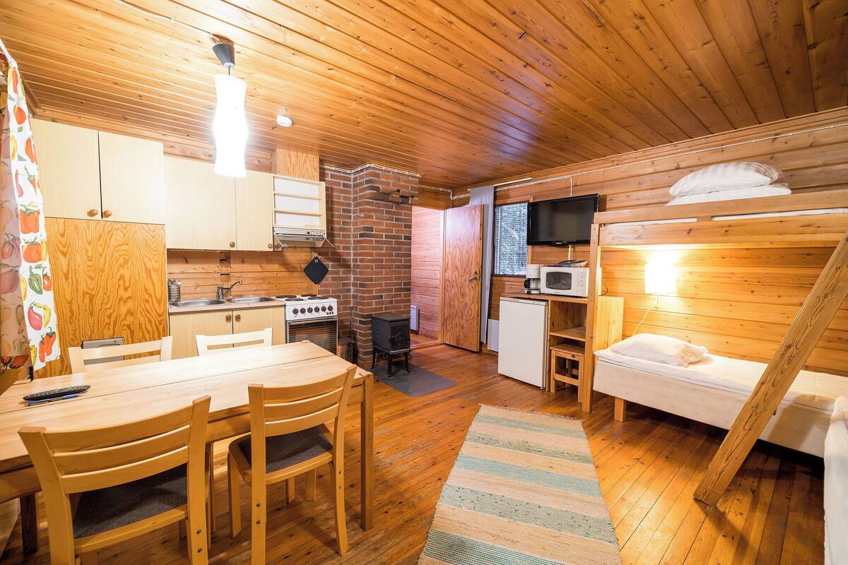 Interior acogedor de una cabaña con una mesa de comedor de madera, sillas, una cocina con estufa y refrigerador, un televisor pequeño y una litera.