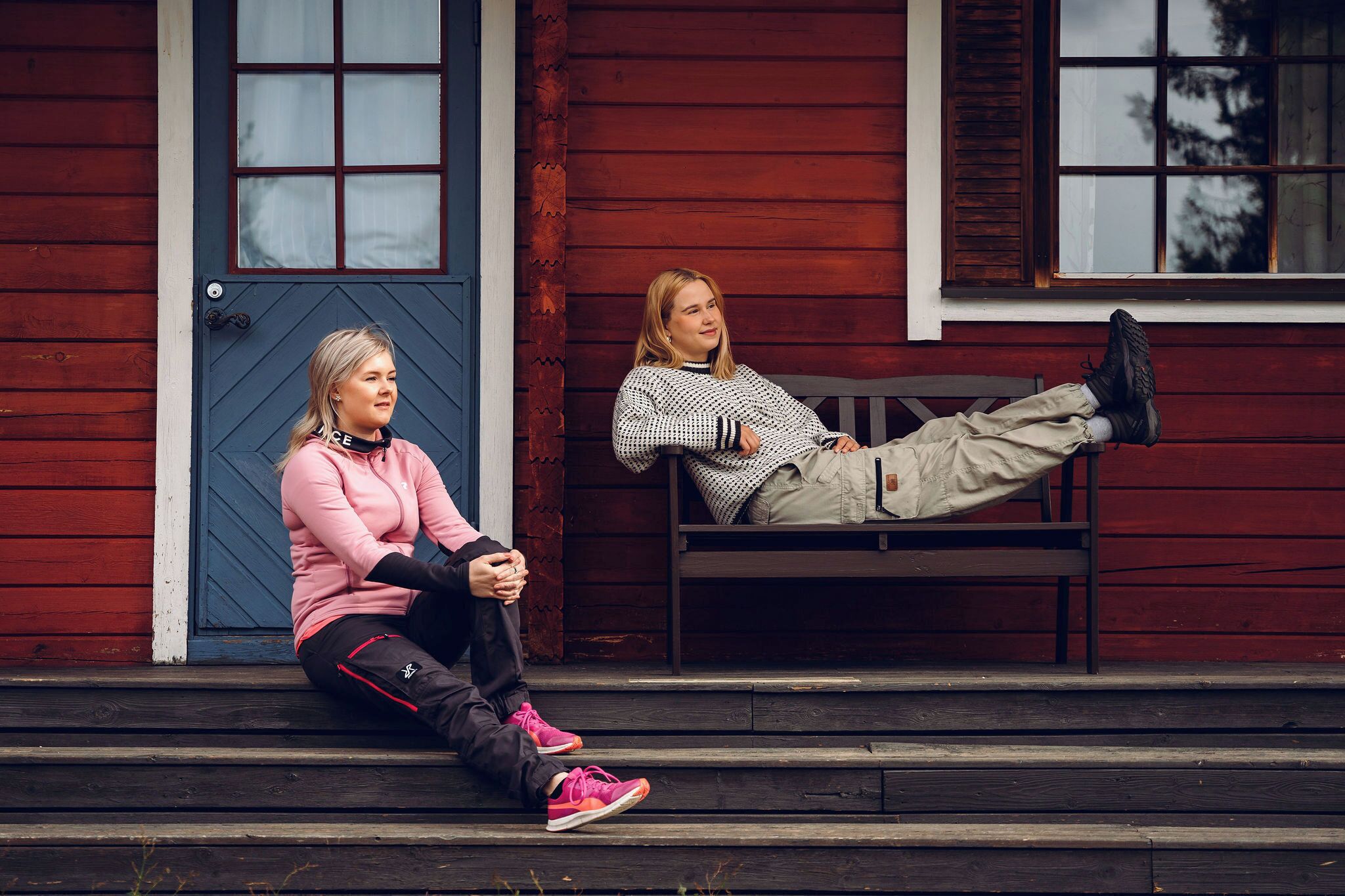 Two people sitting on a wooden bench outside a building with a red exterior and a blue door.
