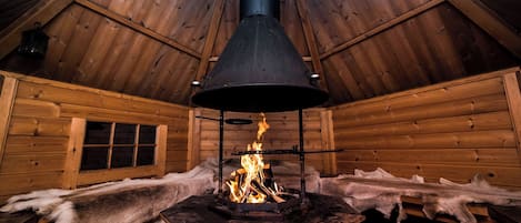 A log cabin interior with a central wood-burning stove, wooden walls, and a sloped ceiling.