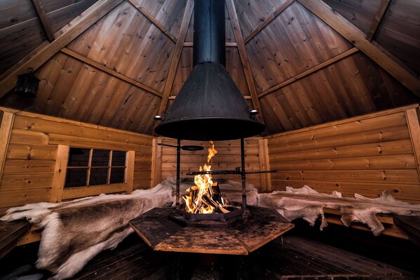 A log cabin interior with a central wood-burning stove, wooden walls, and a sloped ceiling.