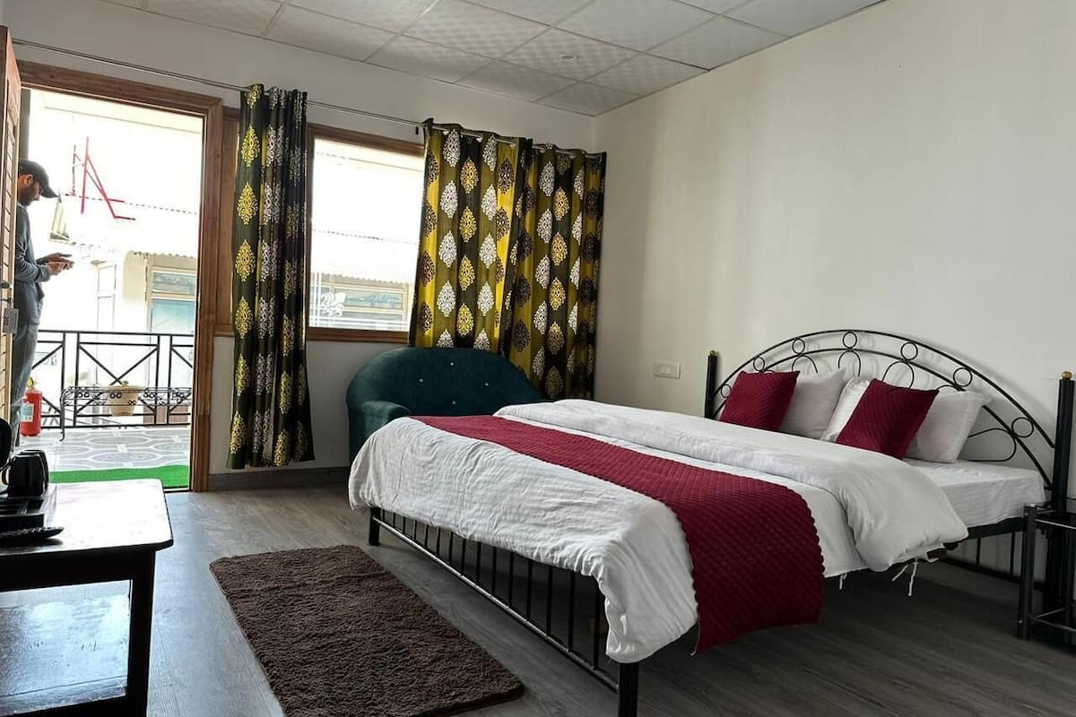 A bedroom with a bed, a desk, and a chair. There is a window with curtains, a door, and a view of the outside.