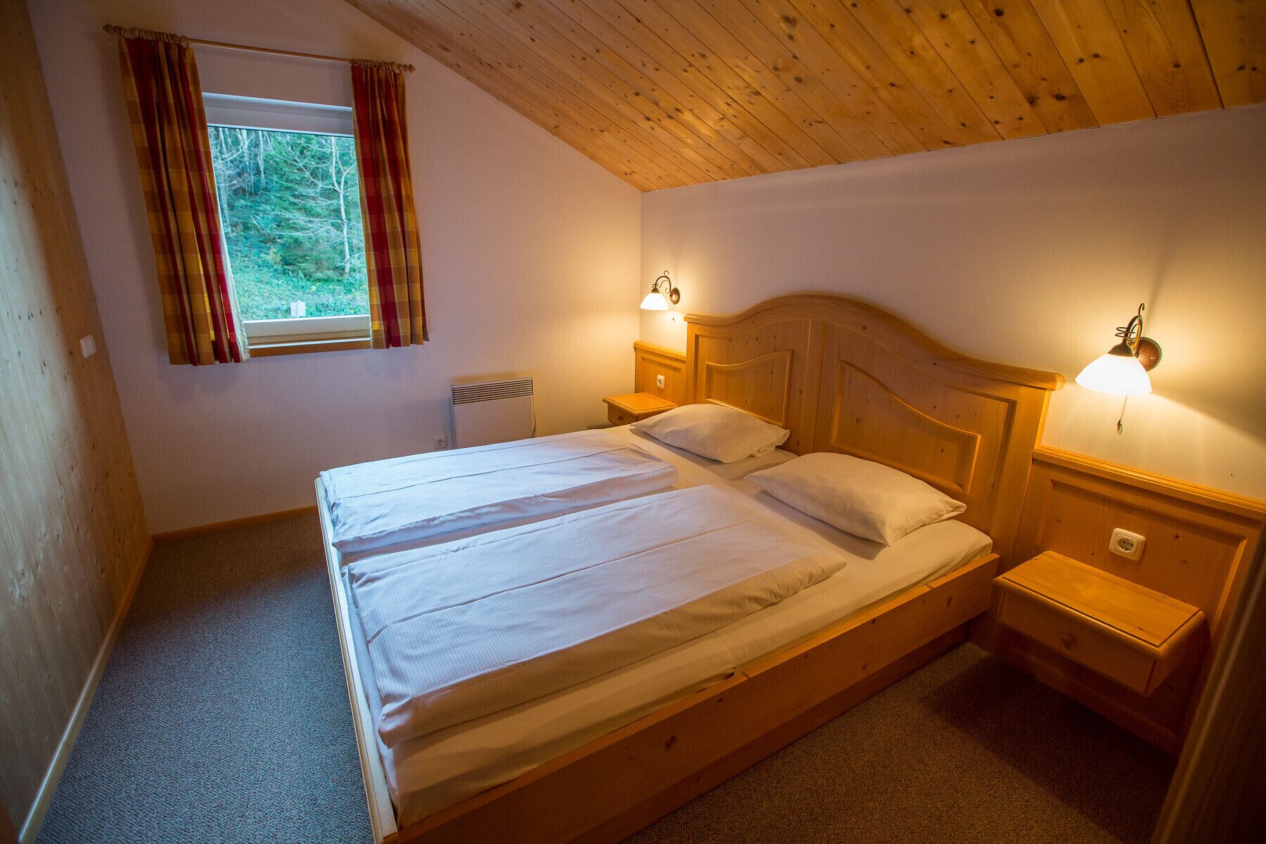 A single-bed room with wooden headboard, two bedside tables with lamps, a window with curtains, and a radiator.