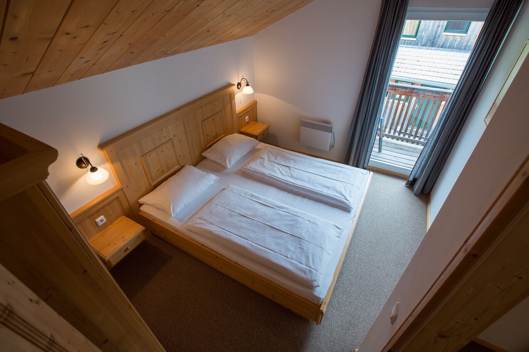 A single bed with a wooden headboard and two bedside tables, each with a lamp. A window with curtains is visible, and a radiator is mounted on the wall.
