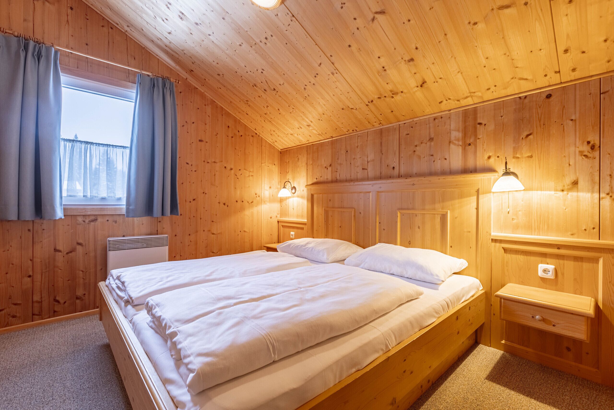 A wooden-paneled room with a double bed, two bedside tables, and a window with curtains.