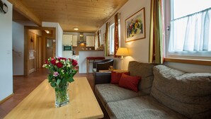A cozy living room with a wooden ceiling, a sofa with red cushions, a table with a vase of flowers, and a kitchen in the background. - Chalet # 56 with 3 BR for up to 6 people (Annaberg)