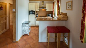 A compact kitchen with white cabinets, a microwave, a toaster, and a small refrigerator. There's a wooden bench with a red cushion, and a window with curtains. - Chalet # 56 with 3 BR for up to 6 people (Annaberg)