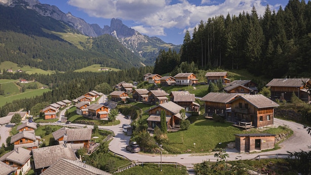 A cluster of wooden cabins with sloped roofs, surrounded by lush greenery and mountains in the background.