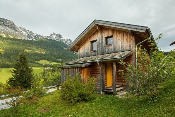 A rustic wooden house with a steep roof, set in a mountainous area with greenery and a road in the background.
