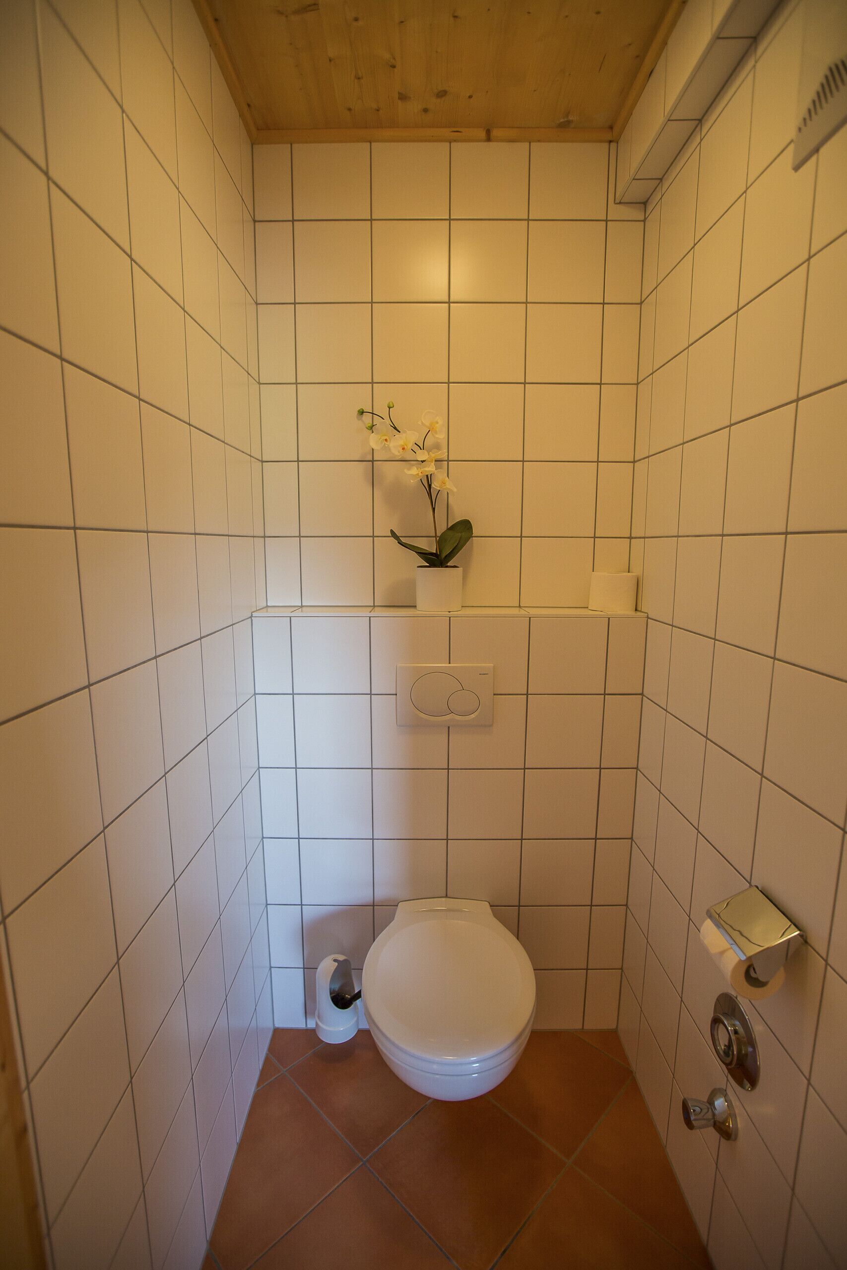 Ein kleines Badezimmer mit Fliesen, einer Toilette, einem Ablagefach mit einem Blumentopf und einem Toilettenpapierhalter.
