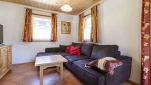 Un salón acogedor con un sofá de cuero oscuro, una mesa de centro de madera y cojines rojos. Hay dos ventanas con cortinas, un techo de madera y un cuadro enmarcado en la pared.
