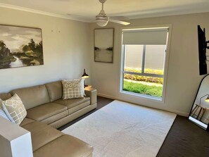 A living room with a beige sofa, a white rug, a ceiling fan, a window with blinds, and a painting on the wall. - Riptide - Pet friendly outside, WiFi (Robe)