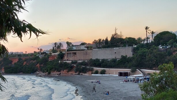 Una playa con olas, una valla de madera y edificios al fondo.