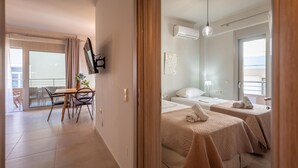 Habitación de hotel con dos camas, zona de comedor con mesa y sillas, balcón con vistas y televisor de pared.