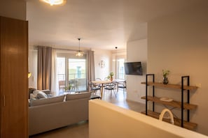 Een moderne woonkamer met een bank, eettafel en stoelen. Er is een tv aan de muur bevestigd en een houten plank met decoratieve objecten.