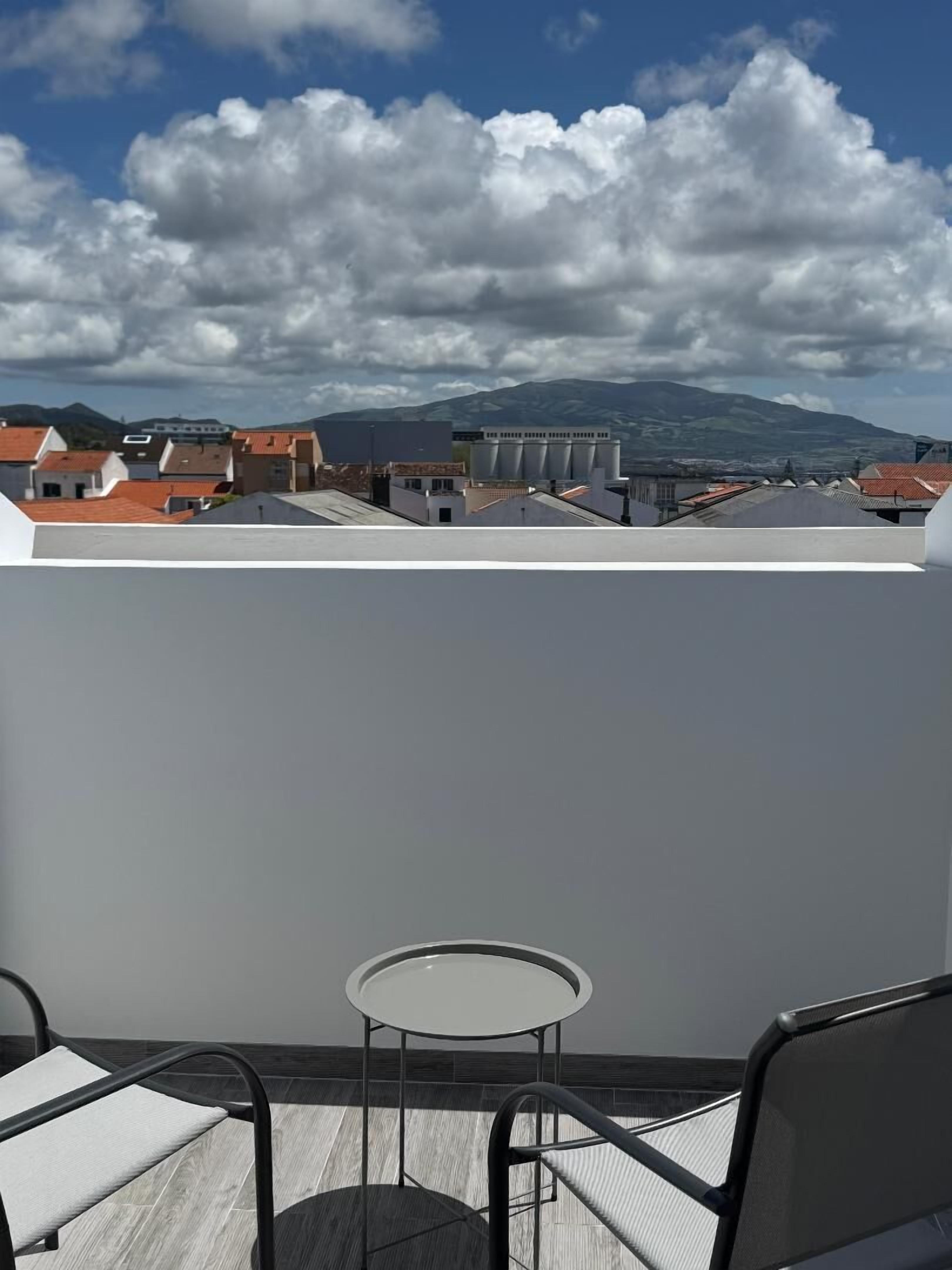 A rooftop terrace with a table and chairs, overlooking a cityscape with buildings and mountains.