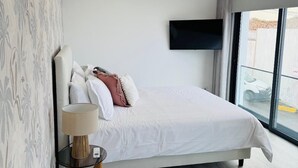 A modern bedroom with a large bed, a flat-screen TV mounted on the wall, a bedside table with a lamp, and a view of the outside through a large window. - Bella Casa Bed & Breakfast (Ponta Delgada)