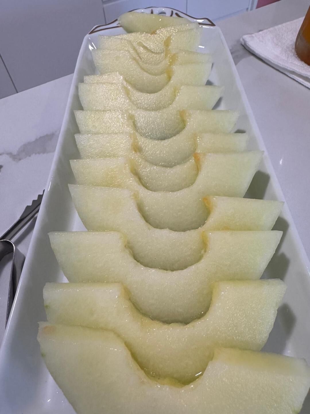 Ein weißer, rechteckiger Teller mit gleichmäßig verteilten, keilförmigen Scheiben eines blassgelben Obstes, vermutlich einer Melone, in einer Reihe angeordnet.