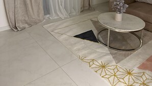 Un salon moderne avec un canapé beige, une table basse ronde ornée d’un vase de fleurs, une grande plante et un tapis à motifs.