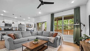 A modern living room with a grey sectional sofa, a wooden coffee table, and a large window with curtains. - Trailside Home | Back 40 + OZ Trails Access (Bella Vista)