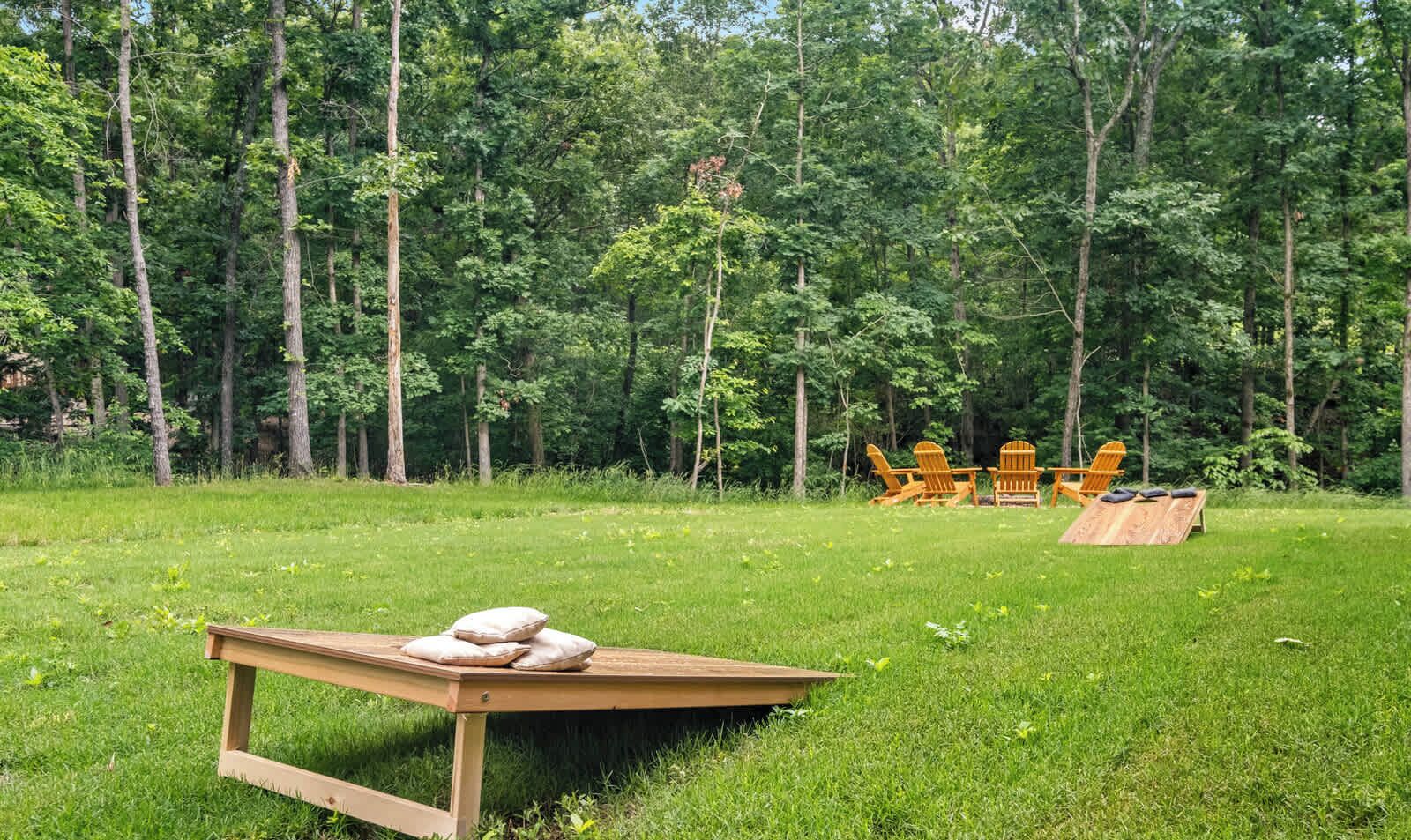 Eine hölzerne Bank mit Kissen, ein Holztisch und Adirondack-Stühle in einer grünen Wiese mit Bäumen im Hintergrund.