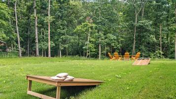 Eine hölzerne Bank mit Kissen, ein Holztisch und Adirondack-Stühle in einer grünen Wiese mit Bäumen im Hintergrund.
