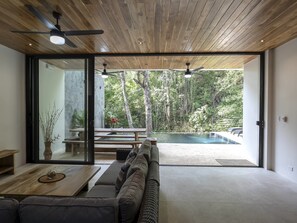 Ein modernes Wohnzimmer mit Holzbalkendecke, großen Schiebeglastüren zum Außenpoolbereich, einem Sofa, einem Couchtisch und einem Deckenventilator.
