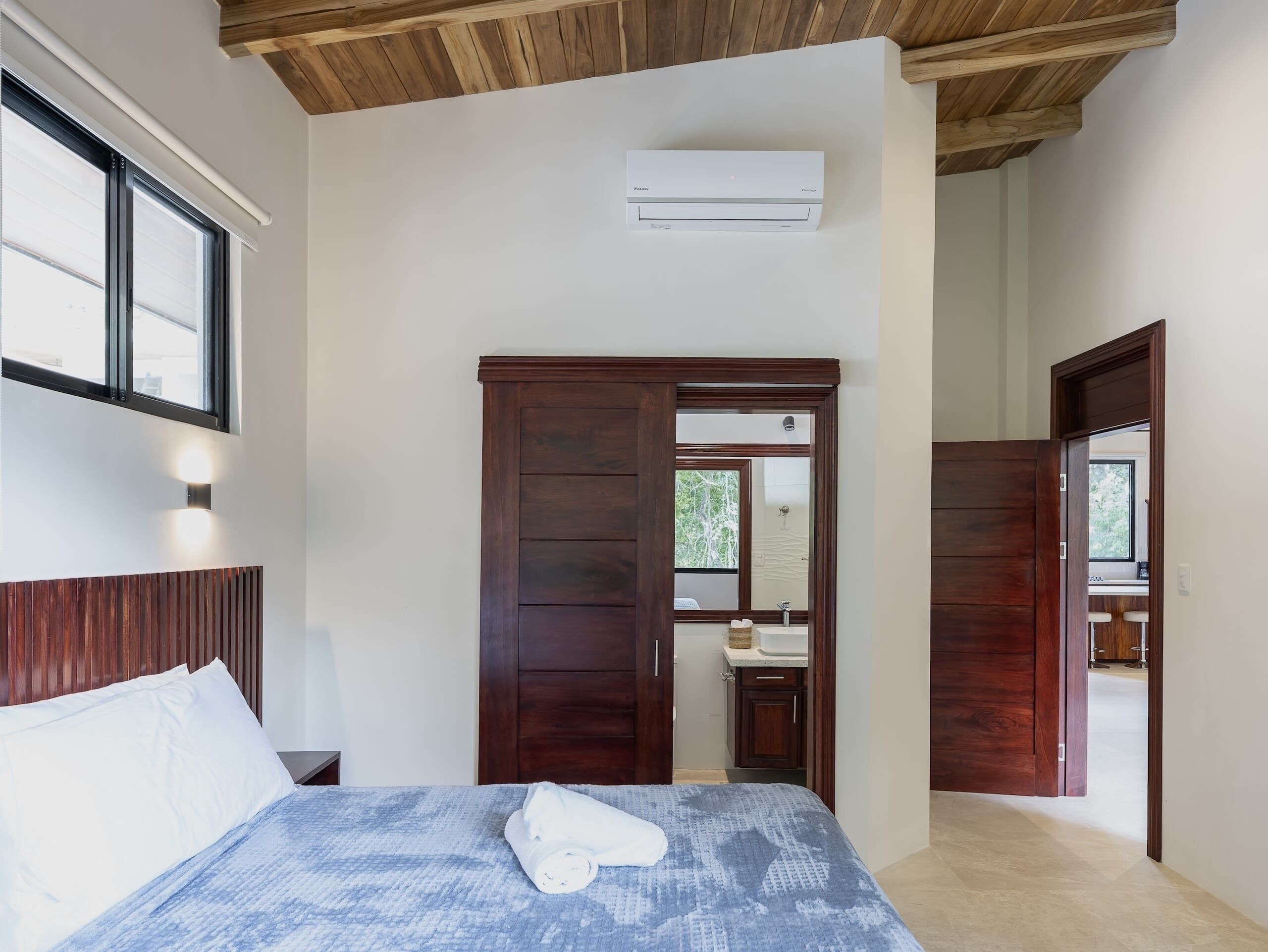 Un dormitorio con cama, armario de madera, baño a la vista por una puerta abierta y un equipo de aire acondicionado en la pared.