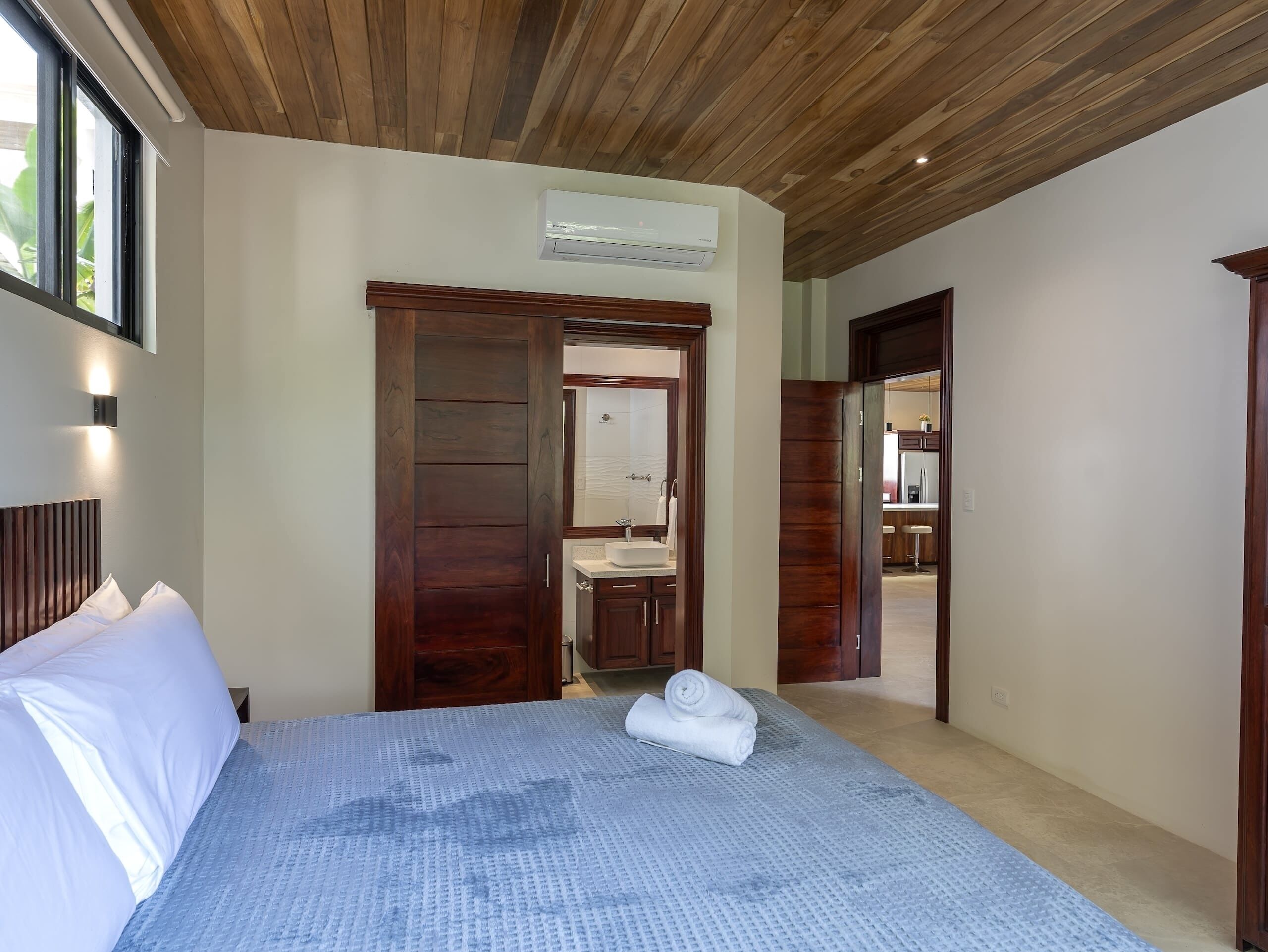 A hotel room with a bed, a wooden ceiling, a bathroom with a sink, and a view of the outside.