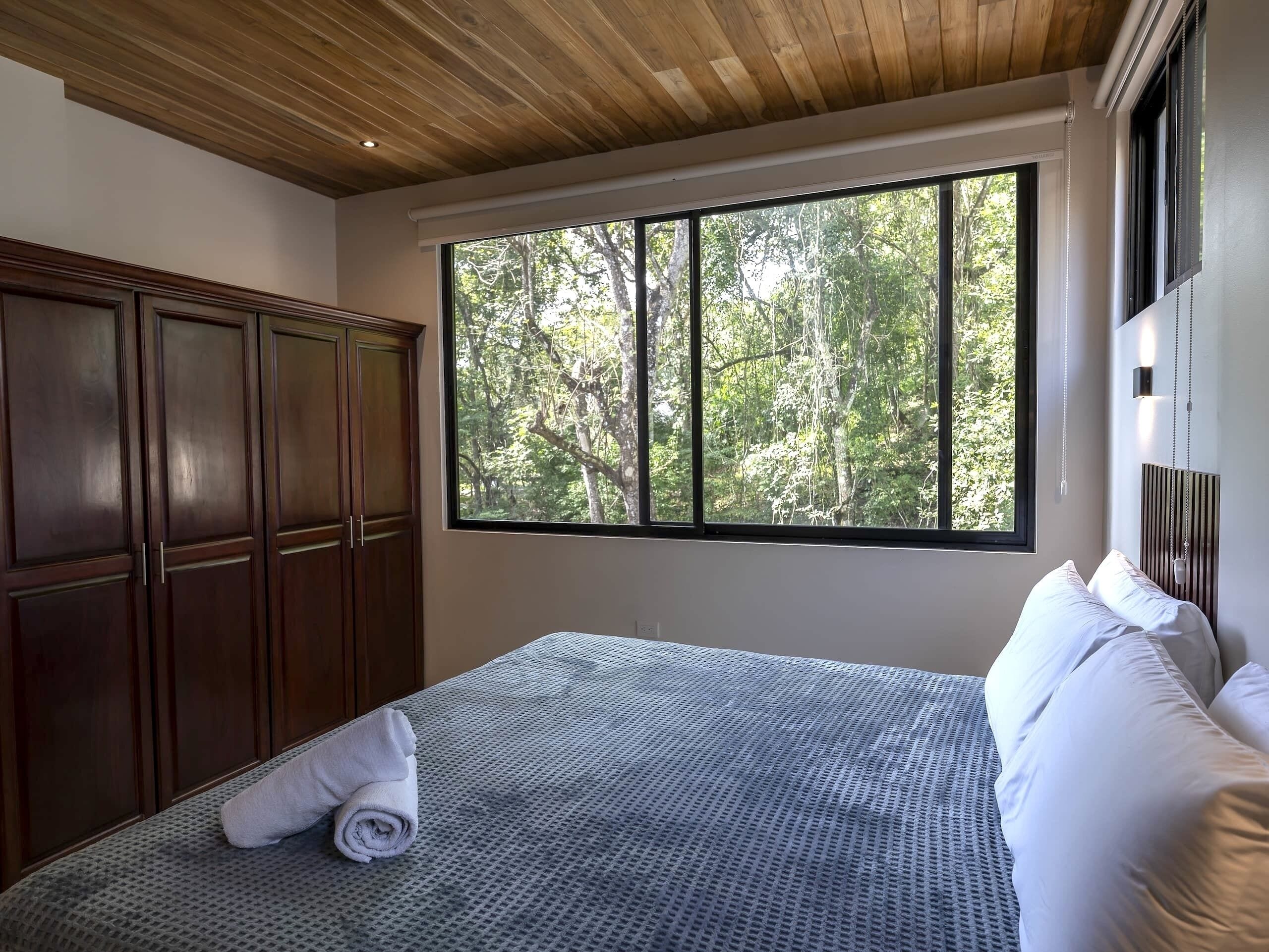 A bedroom with a large window, a bed with white linens, and a wooden wardrobe.
