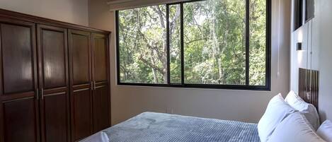 A bedroom with a large window, a bed with white linens, and a wooden wardrobe.