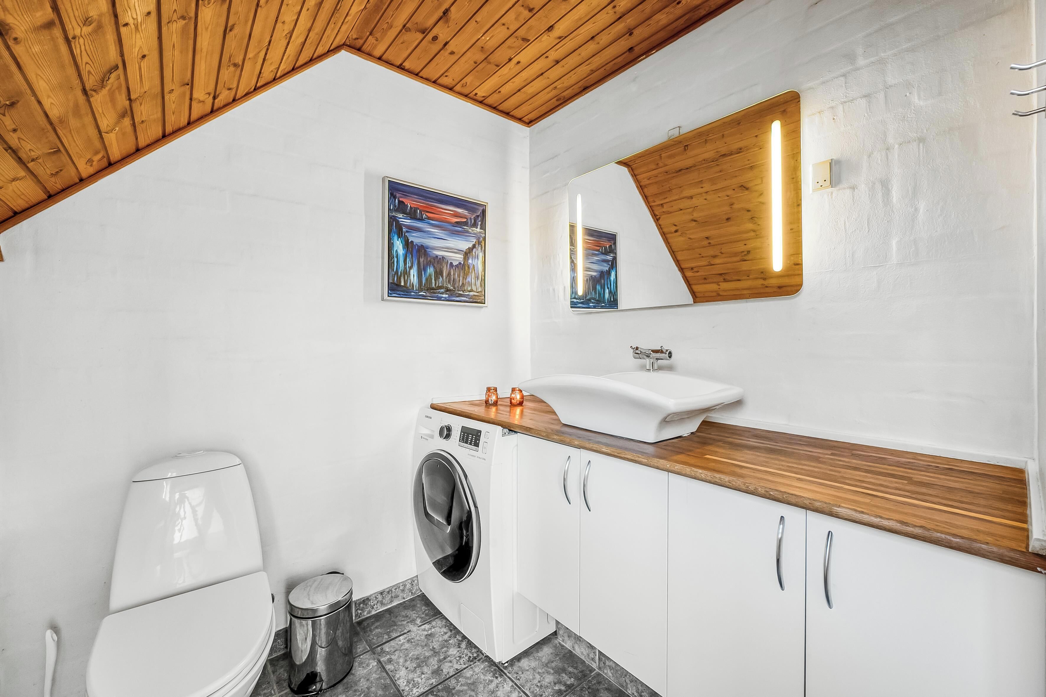Un baño moderno con techo de madera, paredes blancas, lavadora, lavabo con encimera de madera y dos cuadros enmarcados.