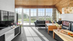 Uma sala de estar moderna com televisão de tela plana, sofás de couro, mesa de centro de madeira e janelas amplas com vista para o exterior.